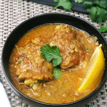 Two coconut chicken thighs in a bowl bathed in lots of coconut curry sauce with a lemon wedge on the side and a cilantro sprig on top.