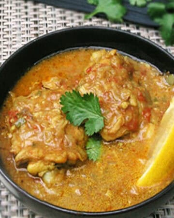 Two coconut chicken thighs in a bowl bathed in lots of coconut curry sauce with a lemon wedge on the side and a cilantro sprig on top.