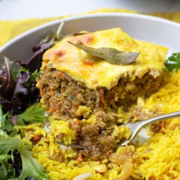 Plate topped with a slice of bobotie, meat casserole with custard on top, yellow rice and salad.