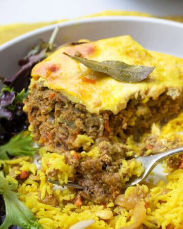 Plate topped with a slice of bobotie, meat casserole with custard on top, yellow rice and salad.