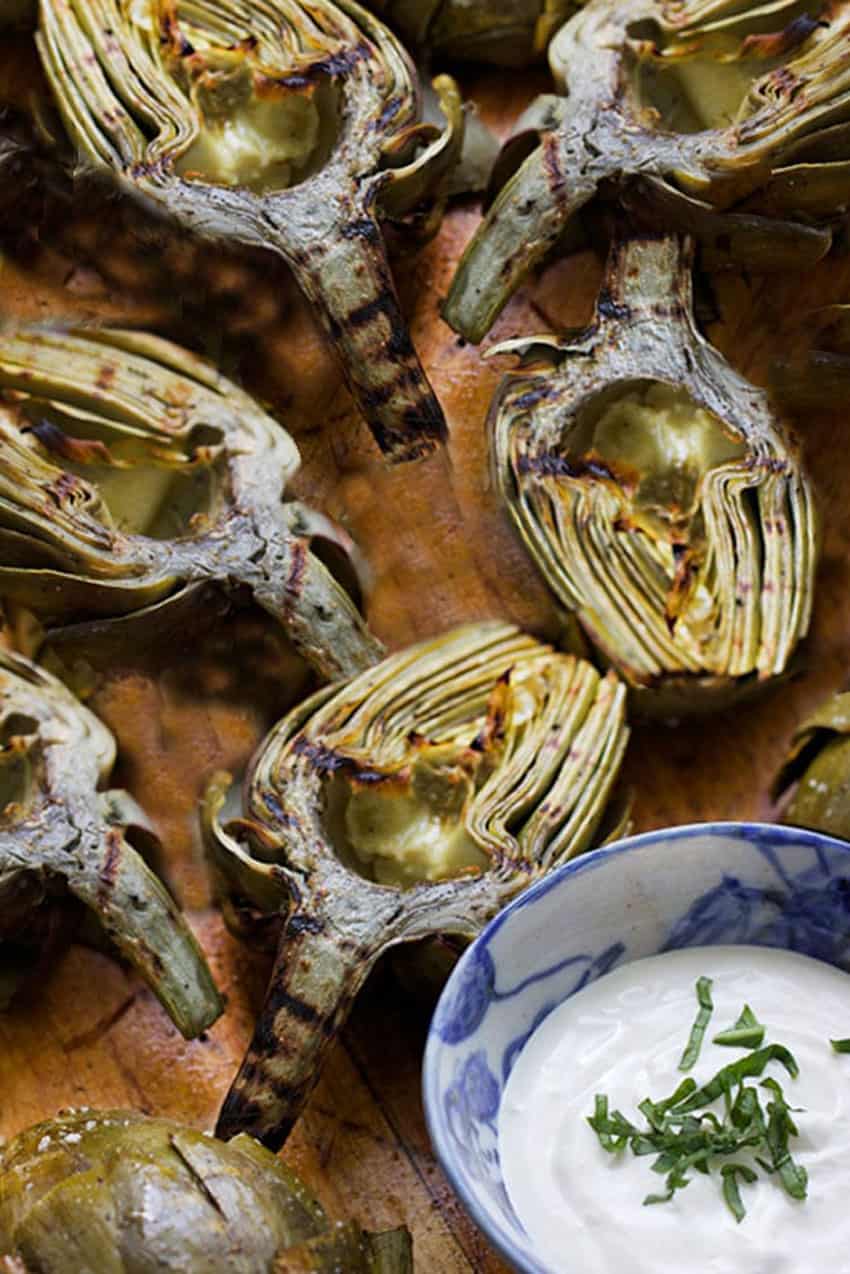 6 halved grilled artichokes on a wooden platter with a bowl of lemon aioli dipping sauce