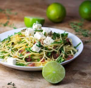 Plate topped with spaghetti twirled with arugula and prosciutto. Cubes of feta on top. Limes in the background and foreground.