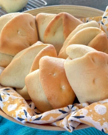 8 coco bread rolls in a basket lined with a flowery cloth napkin.