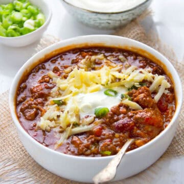 Bowl of chili con carne topped with shredded cheese, sour cream and chopped scallions, a silver spoon in the chili, a small bowl of chopped scallions on the side.