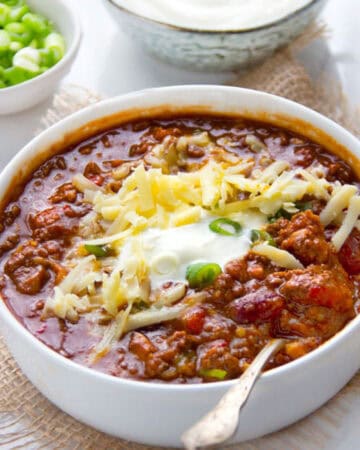 Bowl of chili con carne topped with shredded cheese, sour cream and chopped scallions, a silver spoon in the chili, a small bowl of chopped scallions on the side.