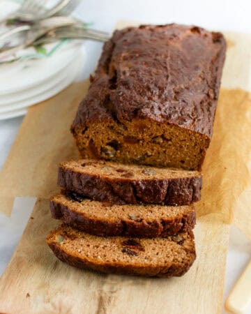 A loaf of pumpkin bread on a wooden board with three slices cut and leaning down from the loaf.