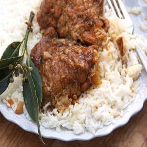 Two piece of Filipino chicken adobo - two braised chicken thighs coated with rich brown adobo sauce - on a bed of white rice, on a plate, with a sprig of bay leaves on the side.