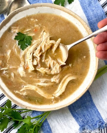 Creamy chicken soup with lots of shredded chicken in a white bowl with a leaf of parsley and a spoon