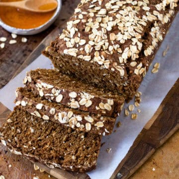 loaf of Irish brown bread on a parchment lined cutting board with three slices cut and a small bowl of honey next to it with a small wooden spoon.