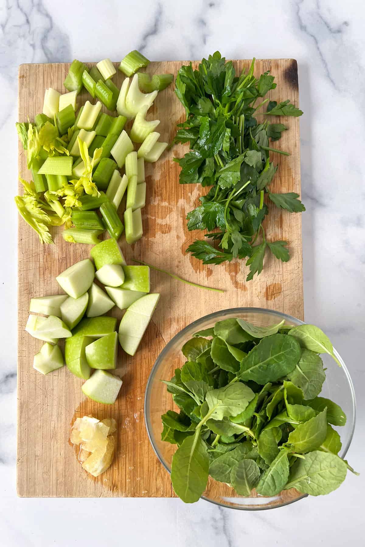 celery, parsley and green apple cut into chunks on a wooden cutting board with a bowl of baby kale leaves.