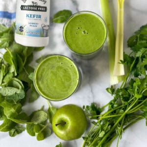 two vibrant green smoothies on a counter surrounded by spears of celery, parsley sprigs, baby kale leaves, a green apple and a bottle of kefir.