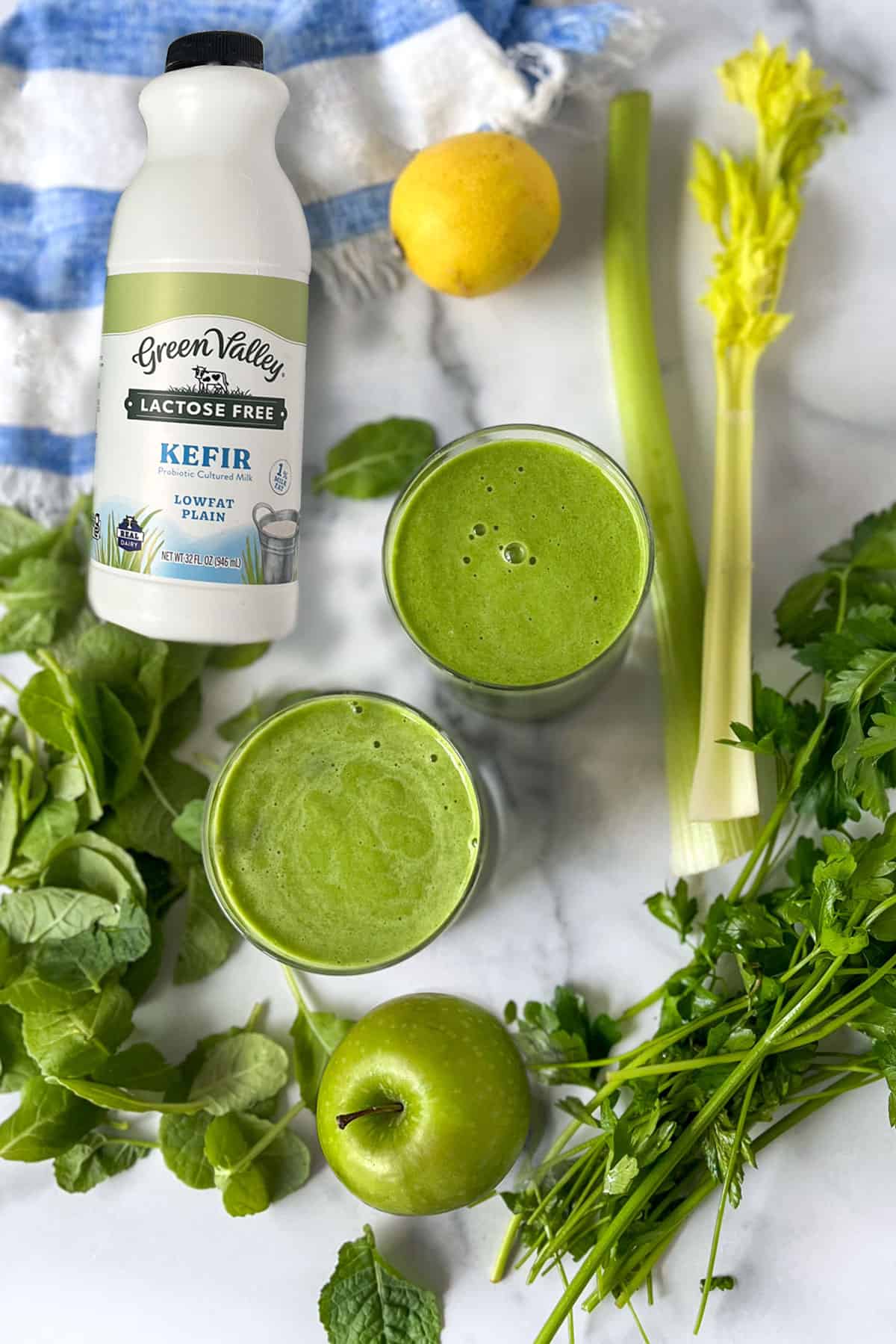 two vibrant green smoothies on a counter surrounded by spears of celery, parsley sprigs, baby kale leaves, a lemon, a green apple and a bottle of kefir.