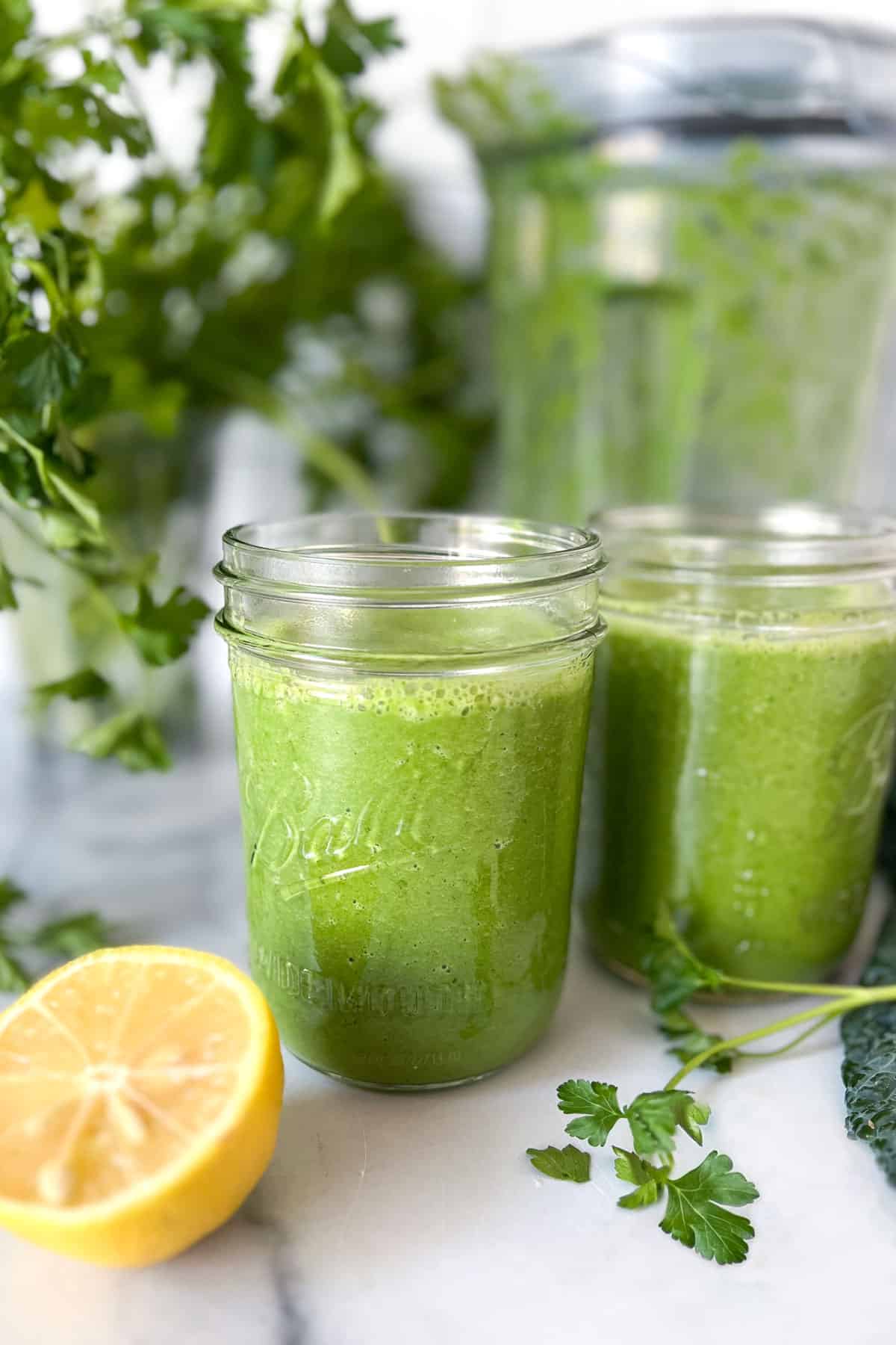 two viviv green smoothies in mason jars, surrounded by kale and parsley and half a lemon.